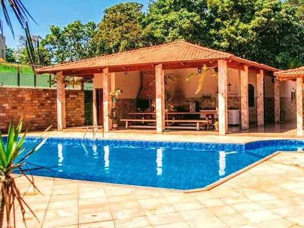 Clínica de Recuperação em Itaúna - MG Piscina azul em piso de pedra clara, diante de um quiosque de telhado cerâmico com mesas de madeira